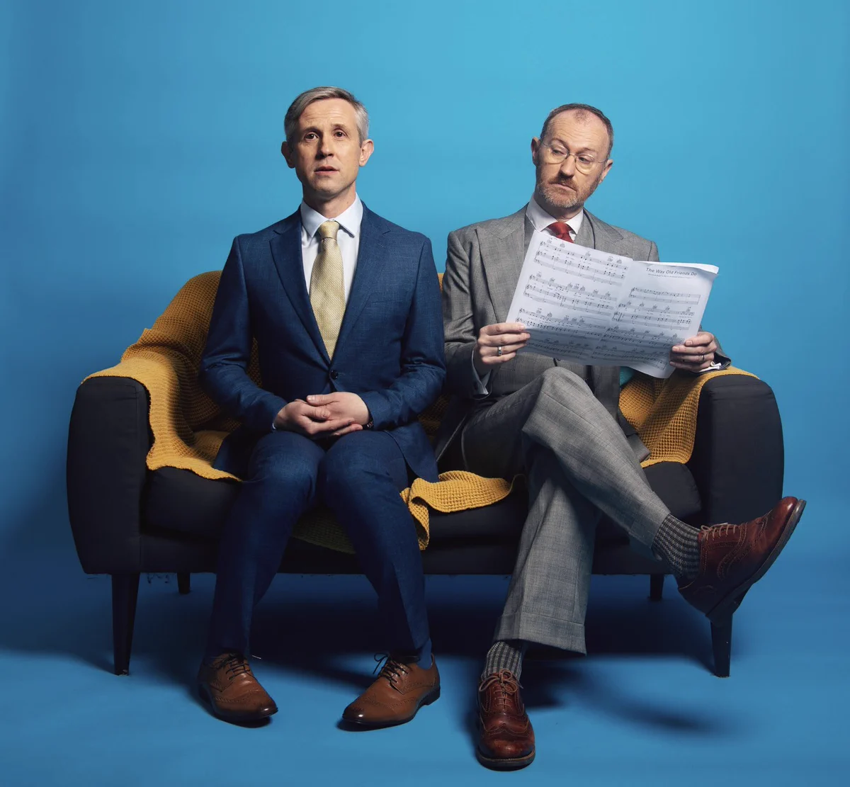 Ian Hallard and Mark Gatiss in suits and ties sit on a sofa, loosely recreating Bengt H Malmqvist’s 1976 photo of ABBA’s Agnetha on a bench looking straight at the camera and Bjorn reading a newspaper. This photo by Darren Bell.