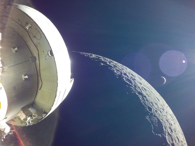 The bottom of the Orion spacecraft shines white in the foreground against the blackness of space. The gray Moon is displayed with visible deep craters in the middle as the distant crescent of Earth glows softly behind.