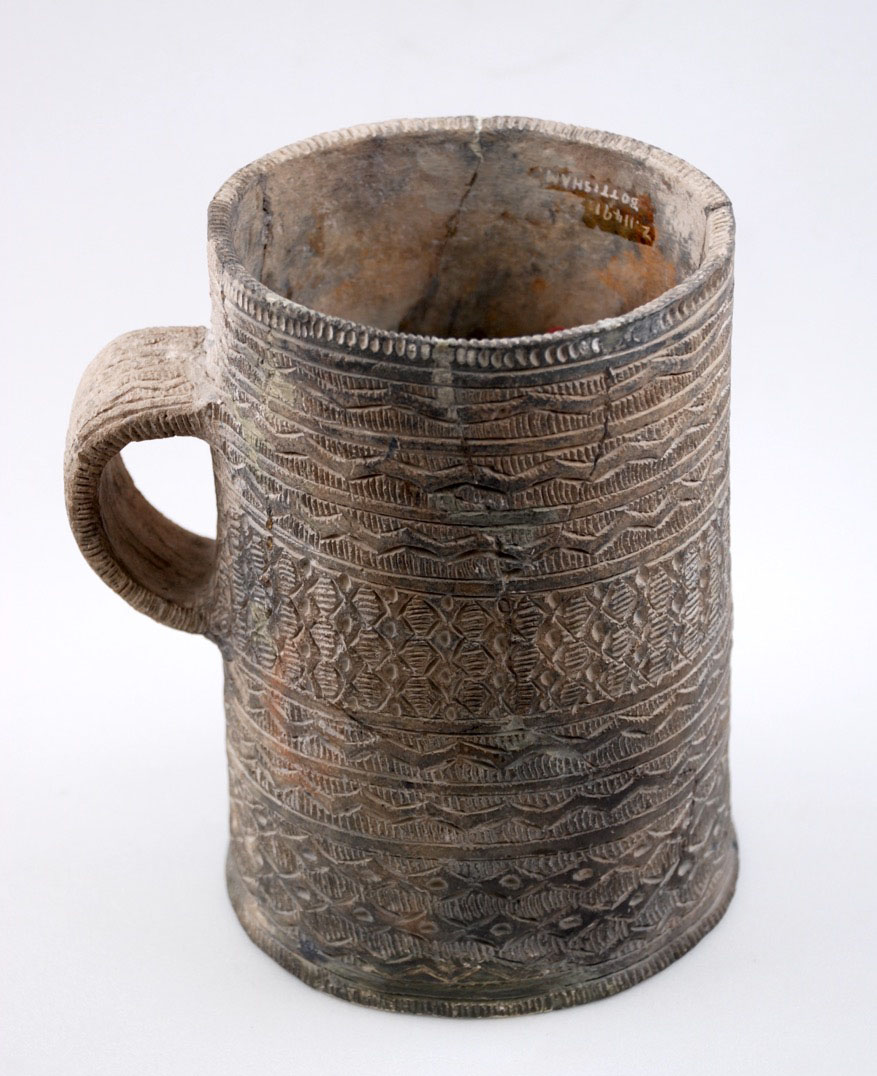 Straight sided, handled beaker. The rim, outer surface and handle are all covered with incised ornament arranged in bands.
