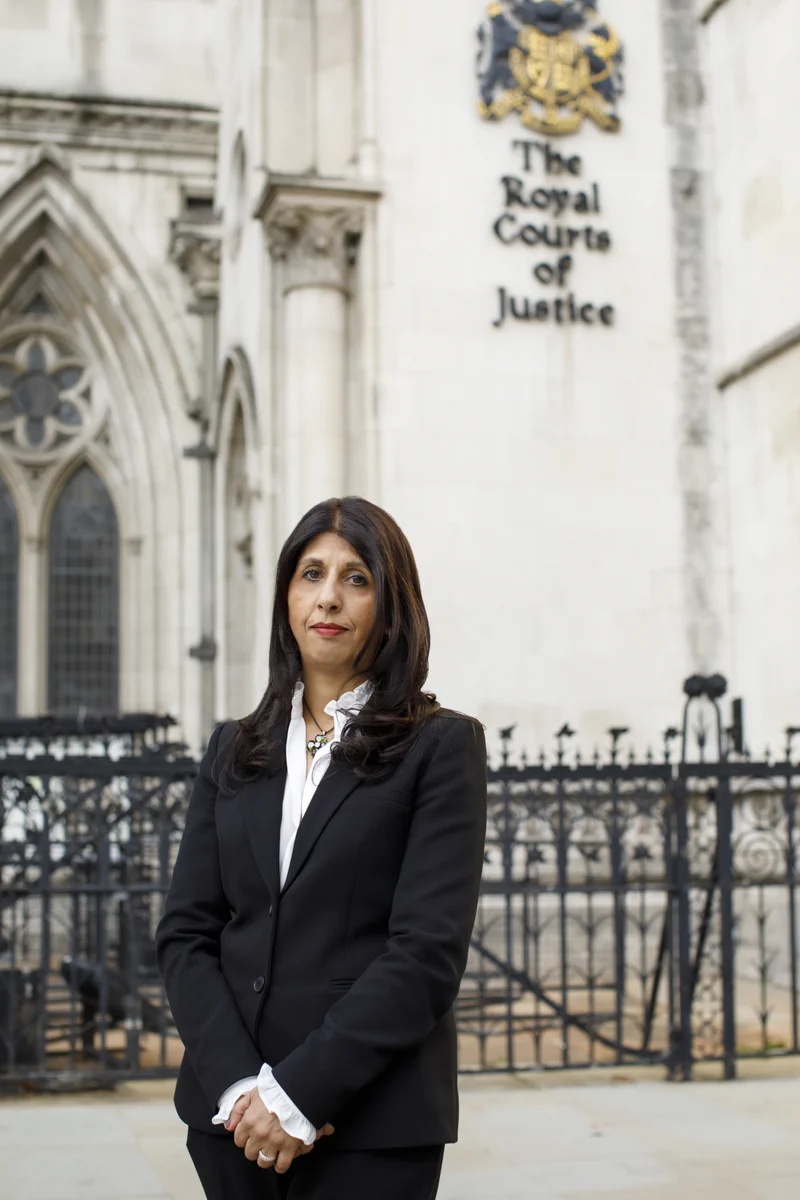 Photo of Lubna Shuja standing outside the Royal Courts of Justice. Asian woman with brown hair, wearing a black blazer and white shirt.