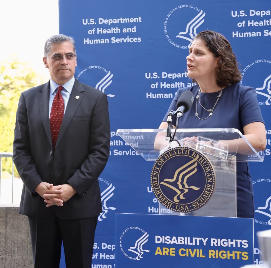 Alison Barkoff speaks a podium with sign that says Disability Rights are Civil Rights. Secretary Becerra looks on, with U.S. Department of Health and Human Services backdrop.