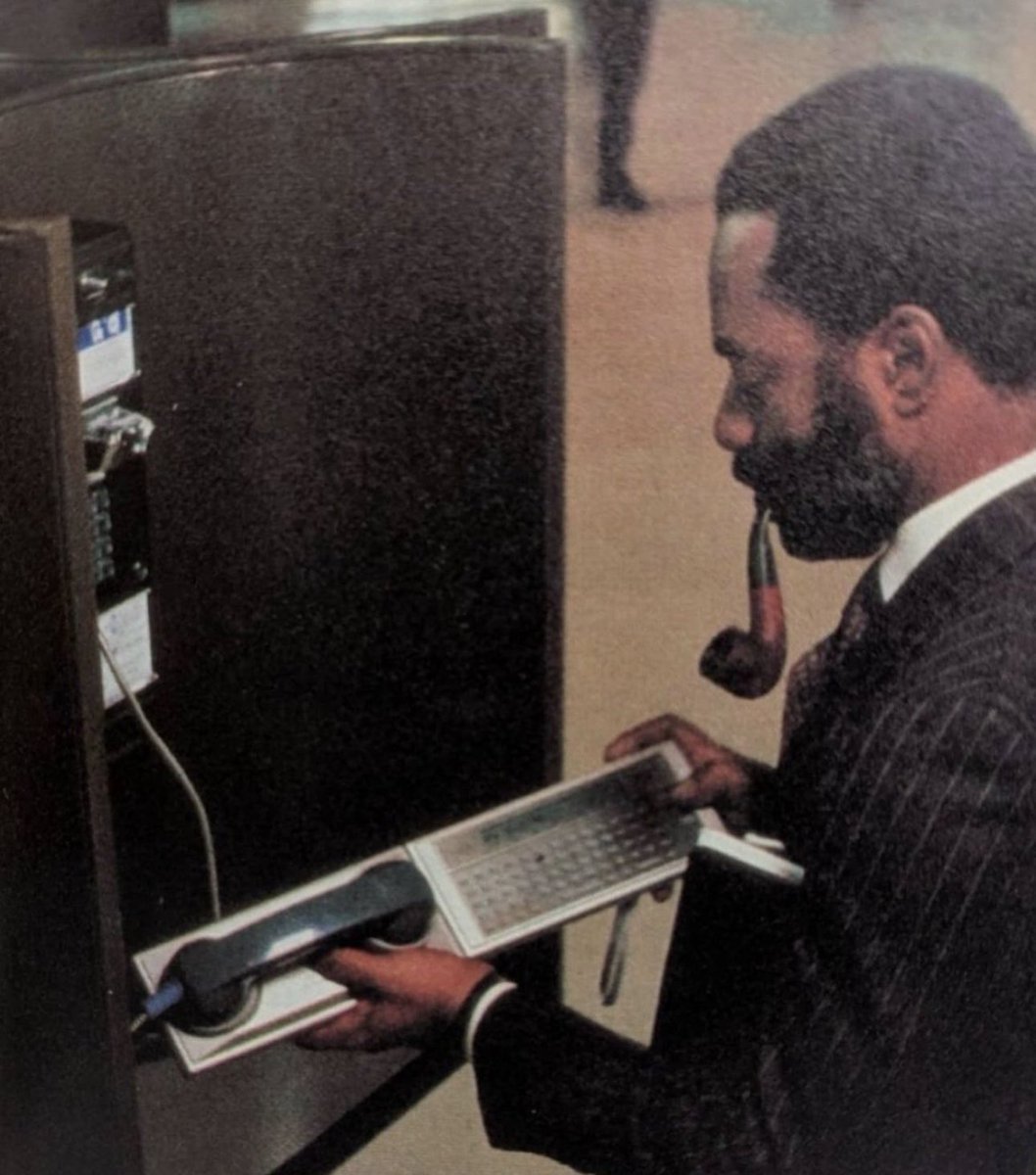 1983, a traveling executive accessing his office email through a Panasonic RL-H1400 handheld computer, using an RL-P4001 Acoustic modem at a public telephone.