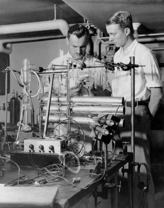 Back and white photo of Compton with graduate student Luis Walter Alvarez standing behind a mass of scientific equipment and looking intently at their work.
