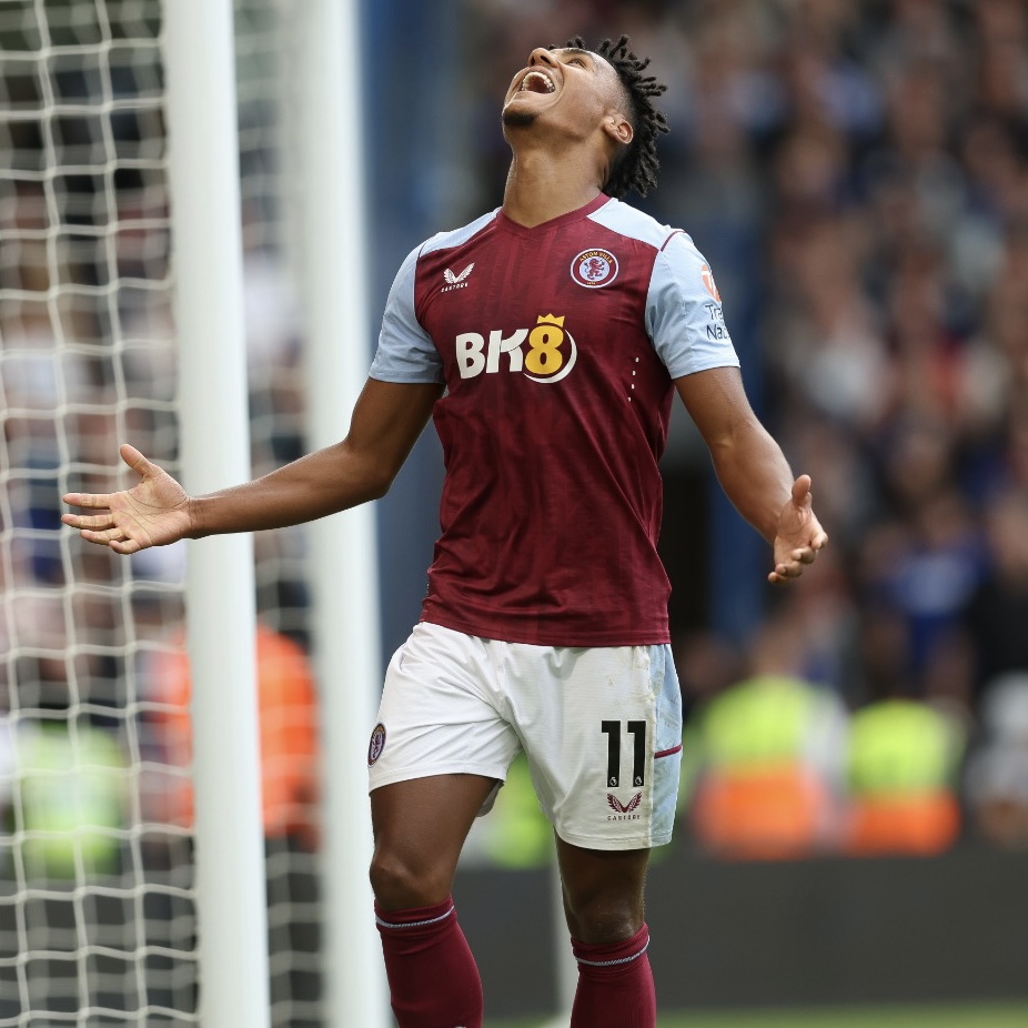 Aston Villa football player Ollie Watkins, wearing the number 11 jersey with 'BK8' sponsorship, stands in front of the goal net with arms outstretched and head tilted back in an emotional celebration.