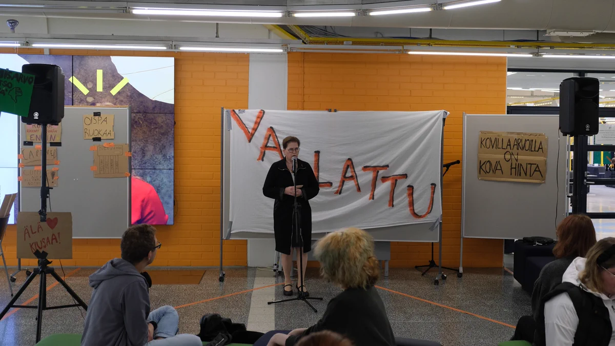 Oulun yliopiston tutkimusrehtori Taina Pihlajaniemi osoittaa Oulun yliopiston johdon tuen yliopistomme Linnanmaan kampuksen vallanneille opiskelijoille, jotka vastustavat Suomen hallituksen suunnitelmia kohdistaa leikkauksia opiskelijoihin. Tutkimusrehtorin takana on lakana, jossa lukee teksti vallattu.