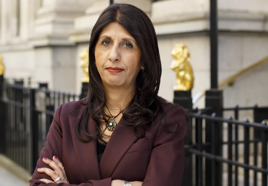 Photo of Lubna Shuja. South Asian woman with brown hair and brown eyes wearing a dark red blazer standing with her arms crossed outside 113 Chancery Lane. 