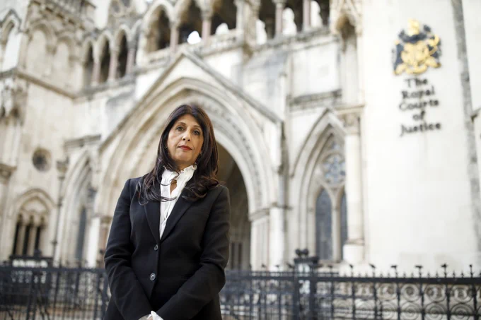 Photo of Lubna Shuja standing outside the Royal Courts of Justice. Asian woman with brown hair, wearing a black blazer and white shirt.  