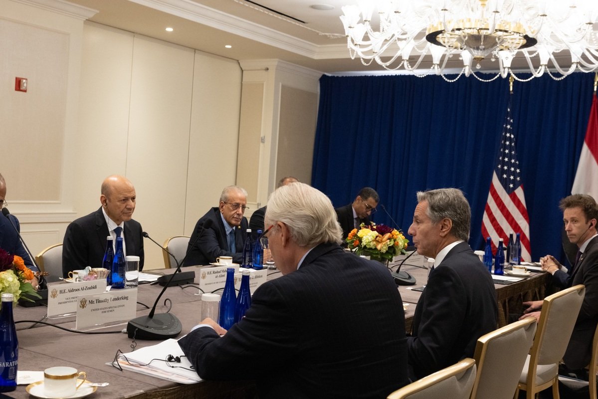 Secretary of State Antony Blinken speaks into a microphone while sitting at a rectangular table across from Yemeni Presidential Leadership Council (PLC) President Rashad al-Alimi. Members of their delegations sit around them. At the end of the table stand large U.S. and Yemeni flags.