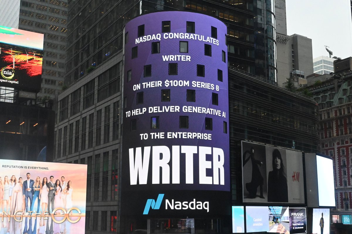The Nasdaq billboard on the side of a New York City building on Times Square. The billboard has a purple background. The caption reads "Nasdaq congratulates Writer on their $100M Series B to help deliver generative AI to the enterprise" Writer logo and Nasdaq logo.