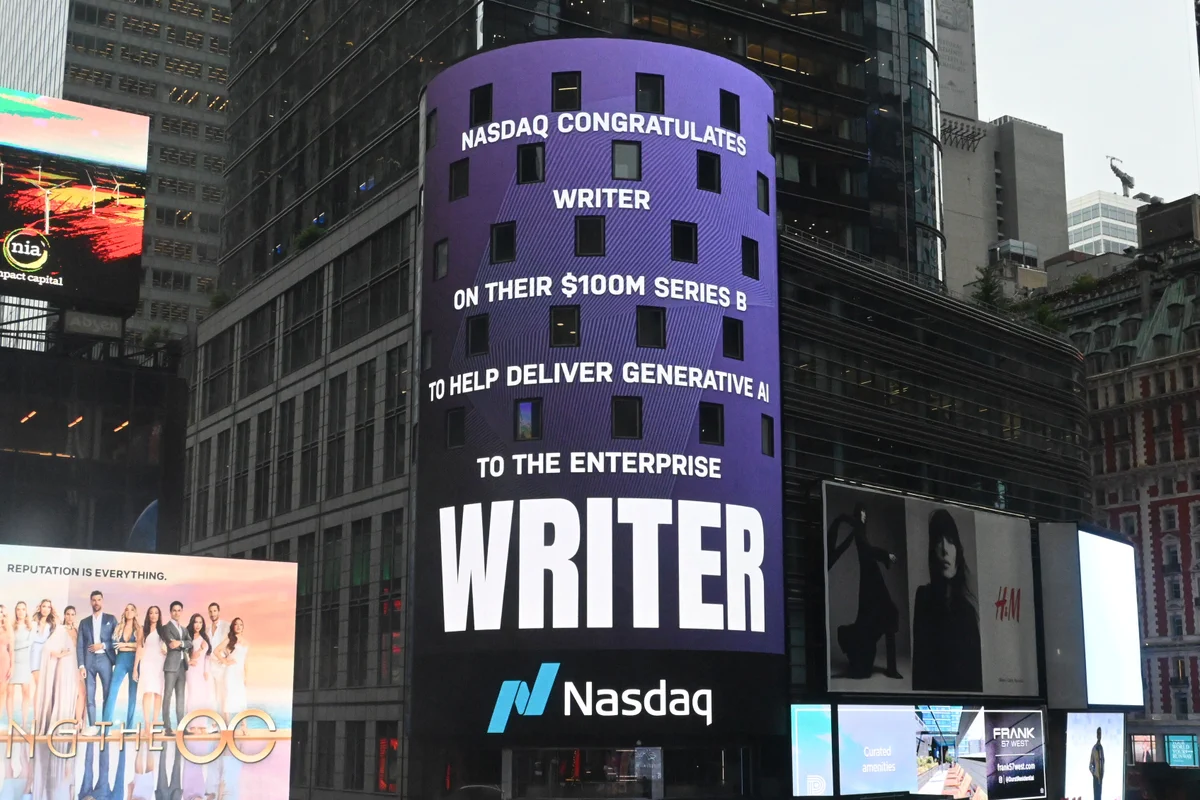 The Nasdaq billboard on the side of a New York City building on Times Square. The billboard has a purple background. The caption reads "Nasdaq congratulates Writer on their $100M Series B to help deliver generative AI to the enterprise" Writer logo and Nasdaq logo.