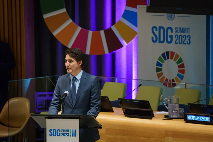 Prime Minister Justin Trudeau is standing at a podium and speaking into a microphone. On the front of the podium, and on a sign behind him, the words “SDG Summit 2023” are written.