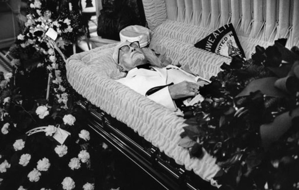 This is an image capturing Colonel Sanders, the founder of KFC, resting in state at the Alumni Memorial Chapel of Southern Seminary in Louisville, Kentucky. 

Sanders was buried in his iconic white suit and a black Western string tie. Approximately 1,000 mourners gathered to pay