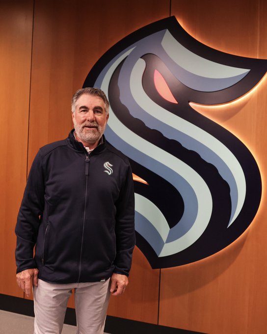 Dave Tippett posing for a photo wearing a kraken jacket in front of a kraken logo