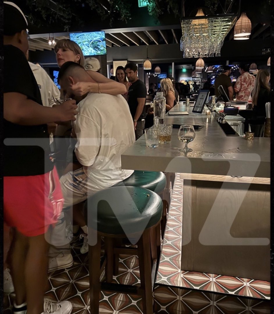 taylor sitting on travis’ lap with her arm around him oh she’s down bad😭