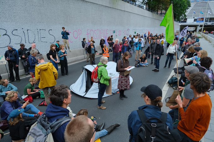 Een zilverkleurige tent staat op de A12. Eromheen zitten en staan demonstranten. Sommige houden vlaggen vast. Op de tunnelwand achter in beeld staat 'Stop fossiele subsidies'. 