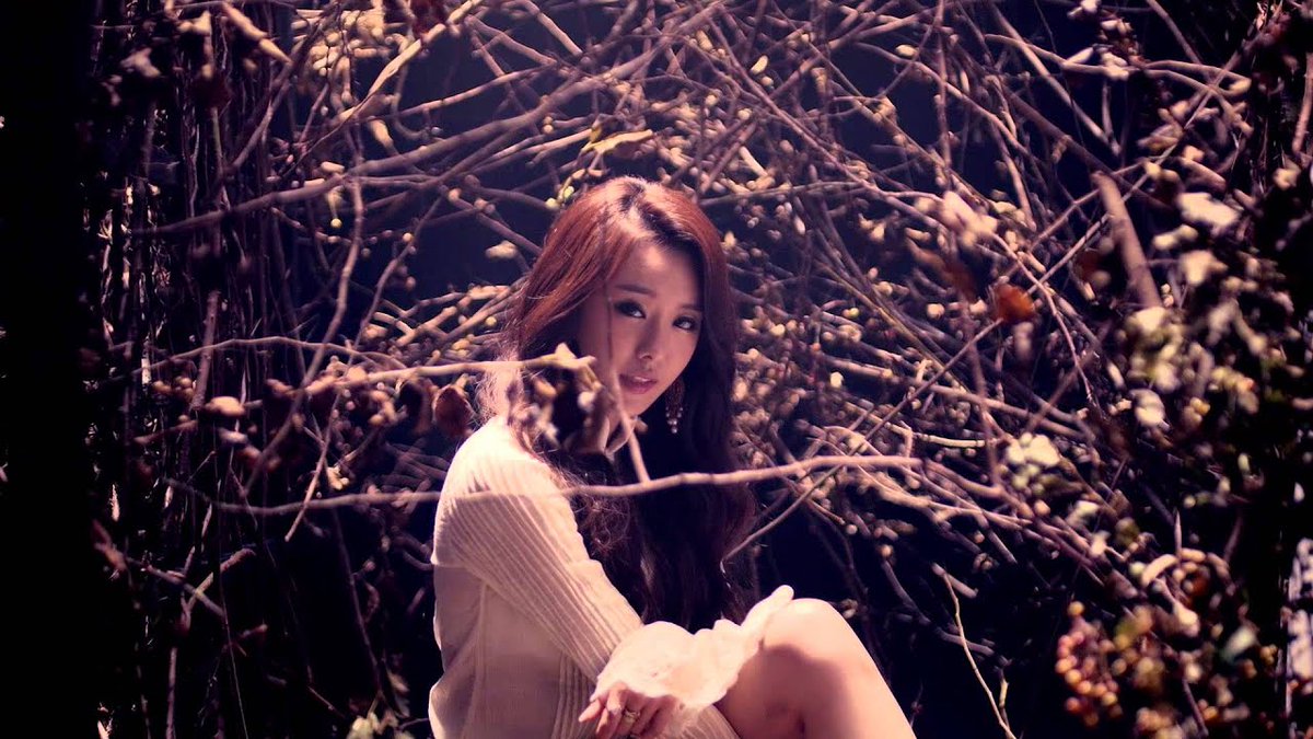 A woman with long brown hair sits in a moody, atmospheric setting surrounded by bare branches and foliage, wearing a cream-colored ribbed sweater.