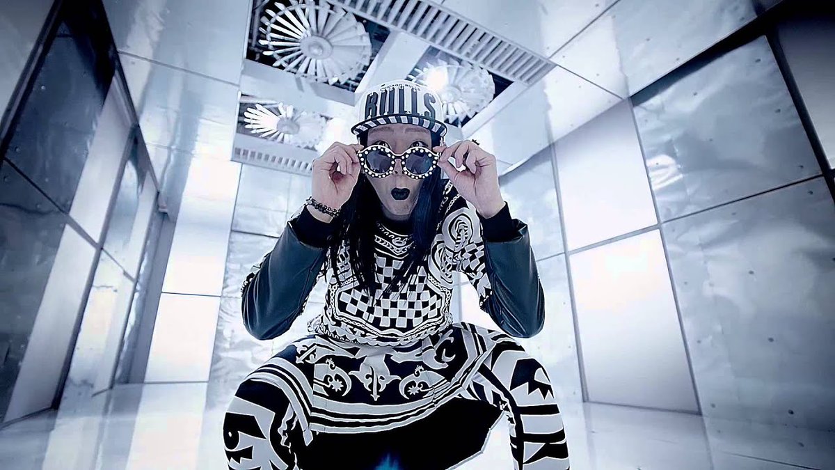 A performer crouches in a black-and-white checkered outfit and 'BULLS' cap, holding decorative sunglasses to their face in a stark, futuristic setting.
