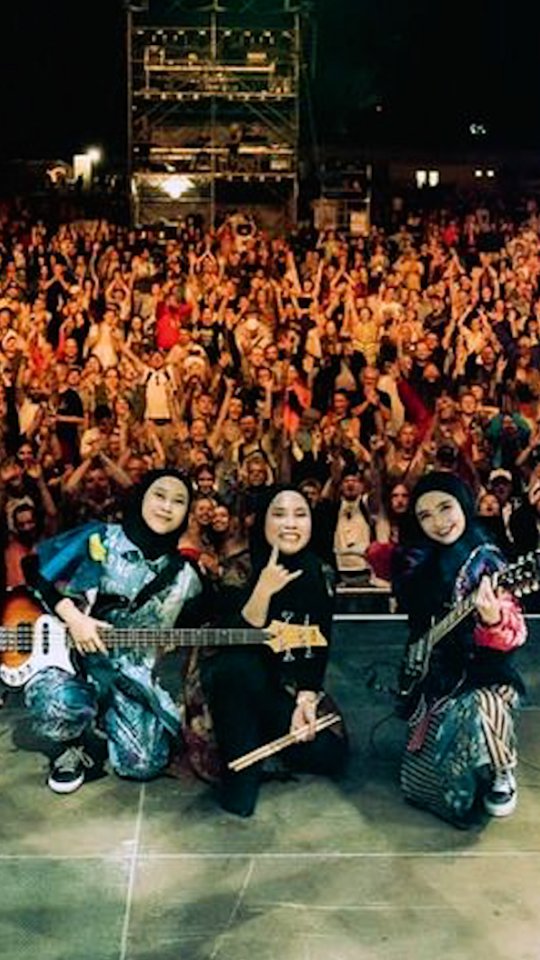 A photo of three female musicians wearing hijabs and holding instruments (a bass guitar and drumsticks) posing on stage in front of a large crowd of concert attendees.