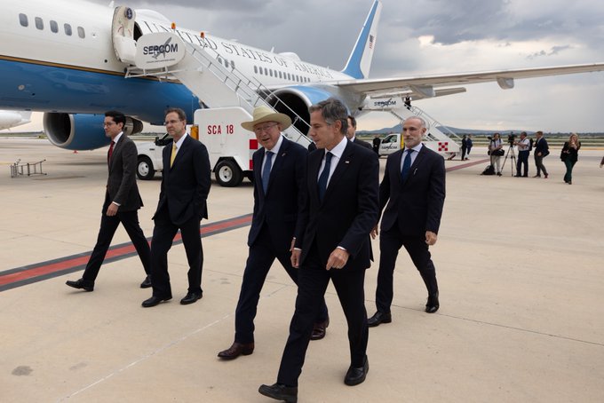 Secretary Blinken speaks to a gentleman while walking across the tarmac upon arrival in Mexico City. Staff and security walk closely behind and his plane sits in the background.