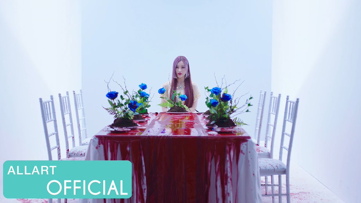A woman with long hair sits at a white-clothed table surrounded by blue rose arrangements in a white room, with the text 'ALLART OFFICIAL' displayed in the corner.