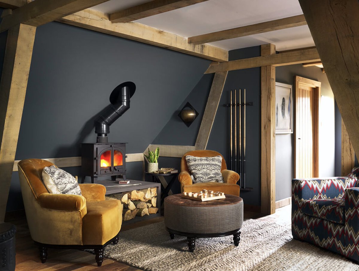Deep armchairs, a wood-burning stove, and a cocktail cabinet feature in this elegant oak barn in the grounds of a country house. The rich colourway of dark grey, mustard, petrol blue, crimson and taupe enhance its cozy.