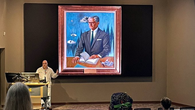George Mashour, MD/PhD, presents in a lecture hall in front of a slide of a surrealist-style portrait of Henry Beecher.