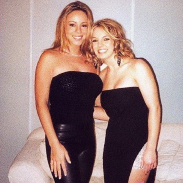 Mariah Carey and Britney Spears back stage. They stand holding each other.