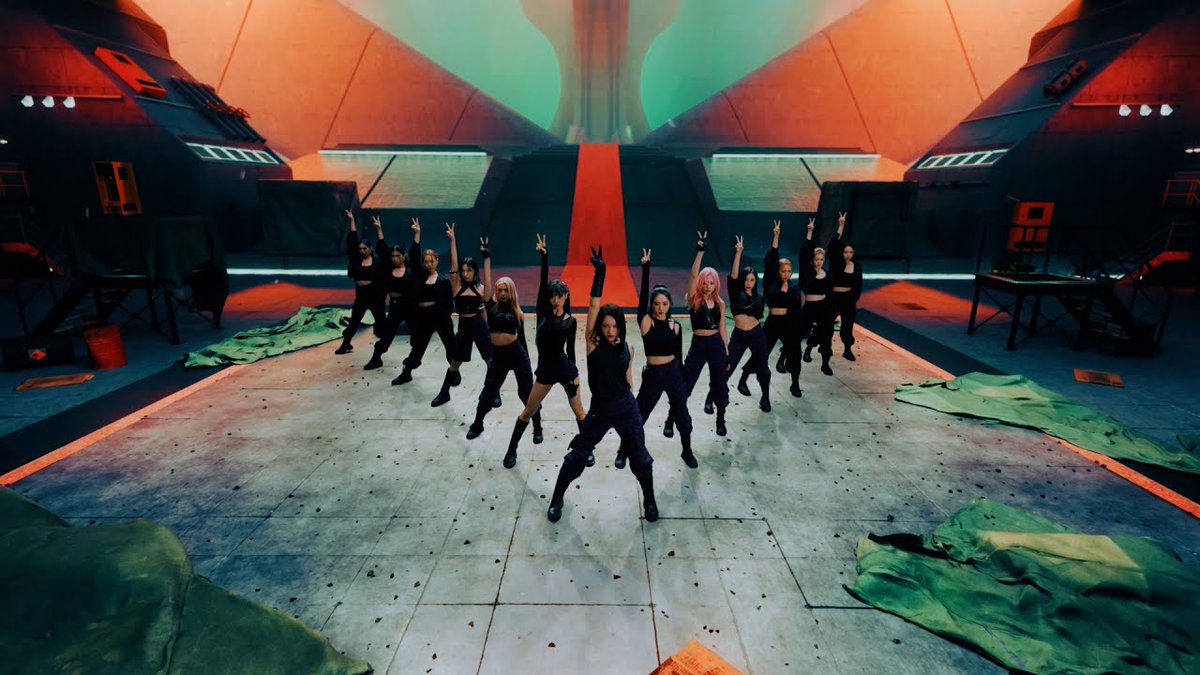 A group of nine female performers strike synchronized dynamic poses in a futuristic hangar-like setting, surrounded by dramatic orange and green lighting and scattered fabric. The image captures the striking choreography from Dreamcatcher's "VISION" music video, marking the 4th anniversary of their 7th Mini Album "Apocalypse: Follow us."