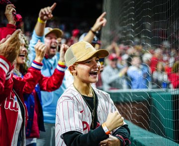 Photo of Liam Castellanos smiling and cheering after Nick Castellanos hit a home run. He's wearing a beige hat, red pinstripes Phillies jersey, and dark shirt.