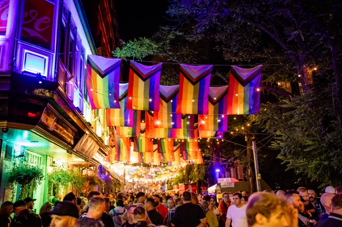 A nighttime photo of Canal Street in Manchester's Gay Village. Canal Street is filled with people celebrating, and looks magical as rainbow lights illuminate the buildings and a canopy of progress pride flags and fairy lights decorate the street.