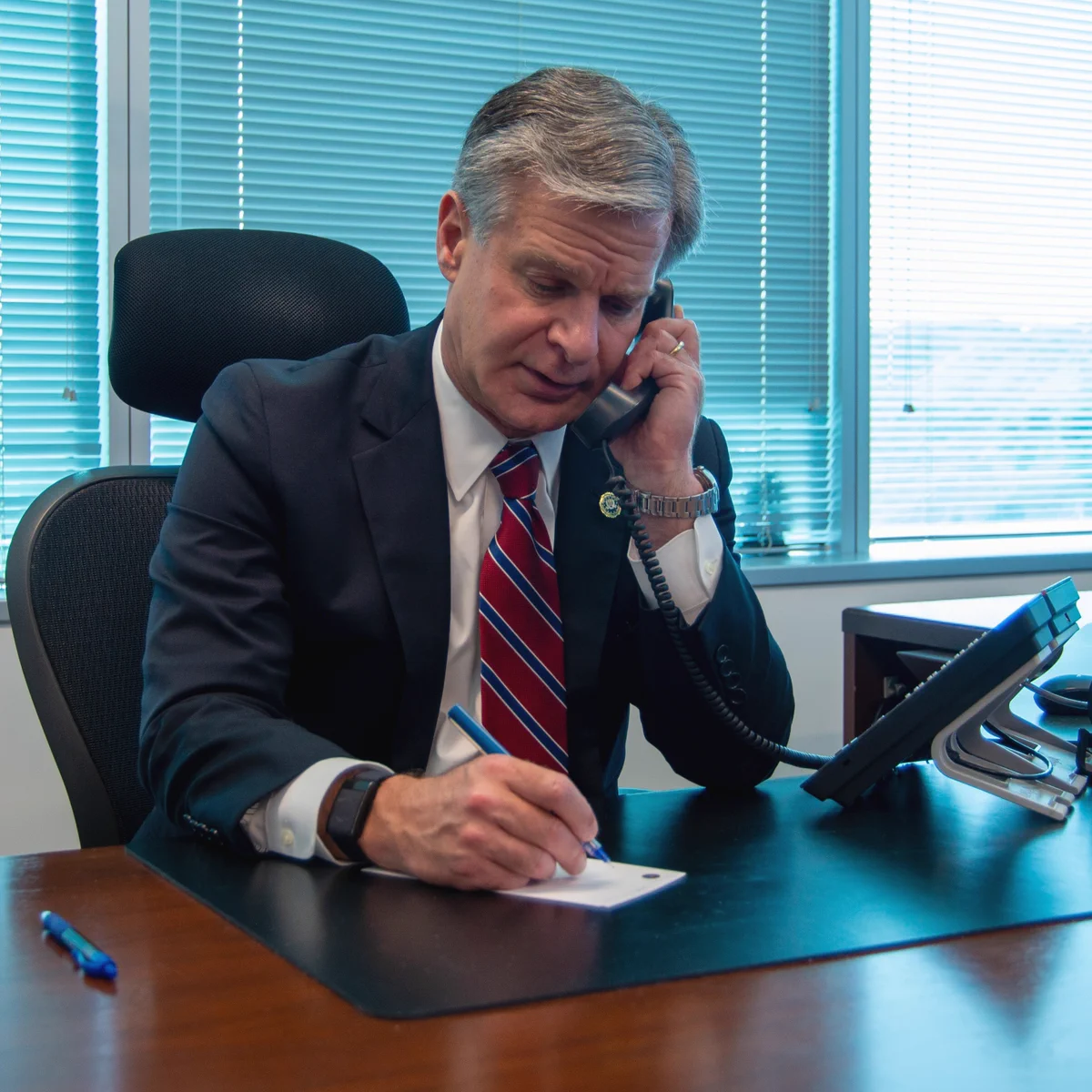 FBI Director Christopher Wray sits in an office, holding a phone to his ear as he participates in the Secure Community Network (SCN) Teleconference.
