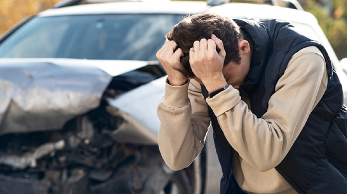 Distressed young man clutching his head with a crashed car behind him.