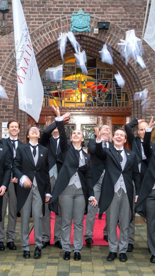 A group of men in formal tailcoats and striped trousers toss their graduation caps into the air in celebration outside a brick building with an arched stained-glass doorway.