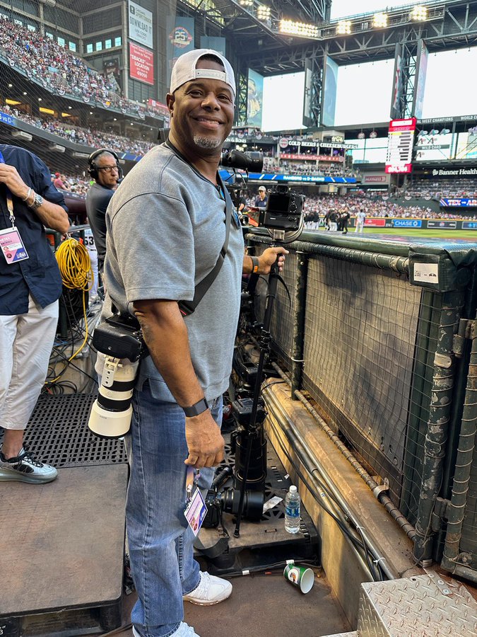 Ken Griffey Jr. wearing gray shirt, jeans and trademark backward cap as he stands in the third base photo well at Chase Field. He holds a camera on a monopod with his left hand with another over his left shoulder.