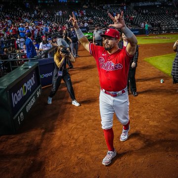 Photo of Kyle Schwarber holding up the love sign after the Phillies win the game to all of the Phillies fans in the crowd. He's wearing the red Phillies jersey and grey pants.