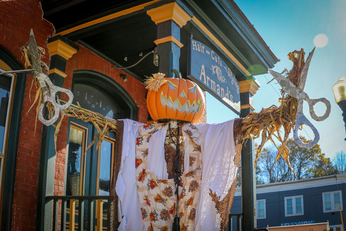 A Halloween decoration with a jack-o-lantern balanced on its shoulders standing in front of a country store on a sunny day.