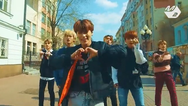 A K-pop boy band performs a choreographed dance on a European-style street, with the lead singer forming a heart shape with his hands while the other members stand behind him.
