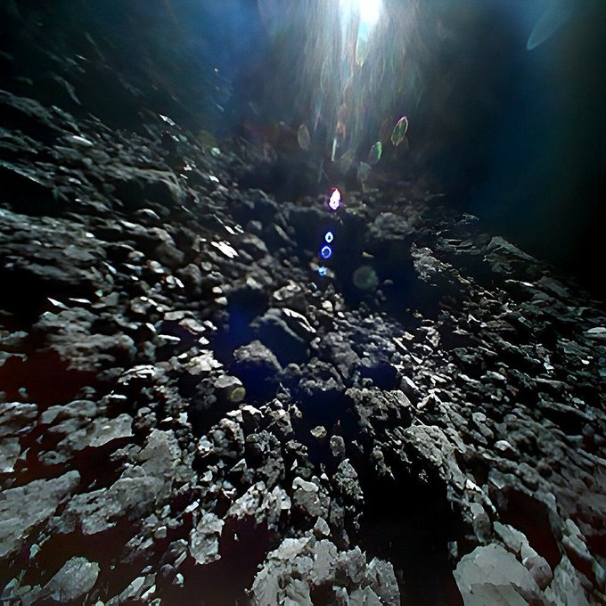The rocky surface of asteroid Ryugu is seen in this video screen grab taken while the small rover Minerva-II-b was bouncing across the surface. A light from the parent spacecraft shines on the surface to illuminate the rocks. The distant rocks are elongated and blurry.