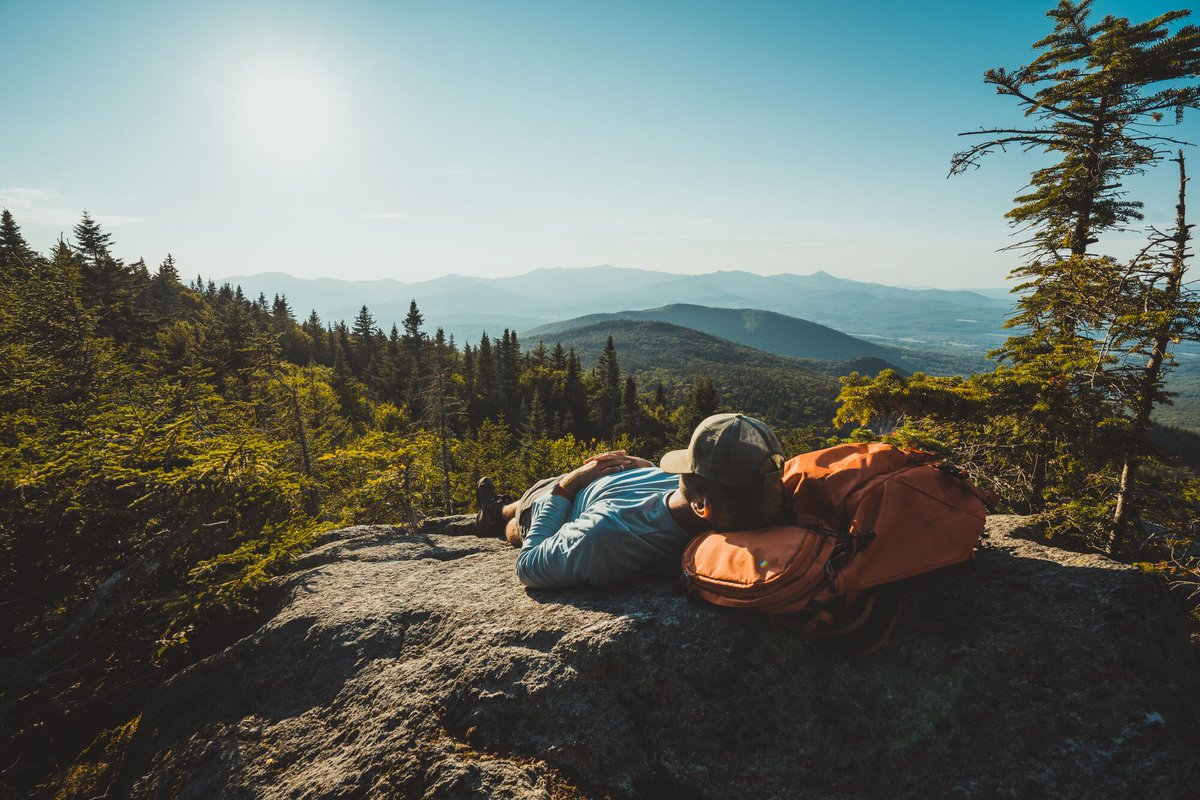 A person at the top of a mountain lying down with their head resting on their backpack and a baseball-style cap pulled down over their eyes. Mountain peaks stretch ahead under a blue sky in the distance.