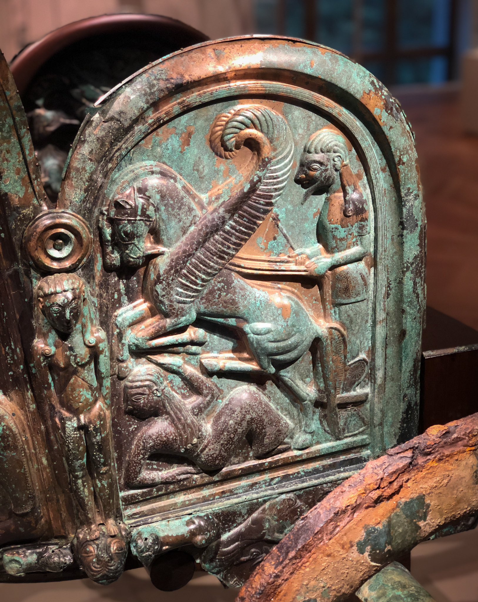 The left side panel of the chariot, depicting the apotheosis of dead Achilles, ascending to the heavens in a chariot that's nearly identical to this one, pulled by two winged horses. A woman has fallen to the ground under the horses, her hand up in defense, her head turned away from the sight. The panels all have a green patina, with polished golden bronze showing through.

Photo: my own.