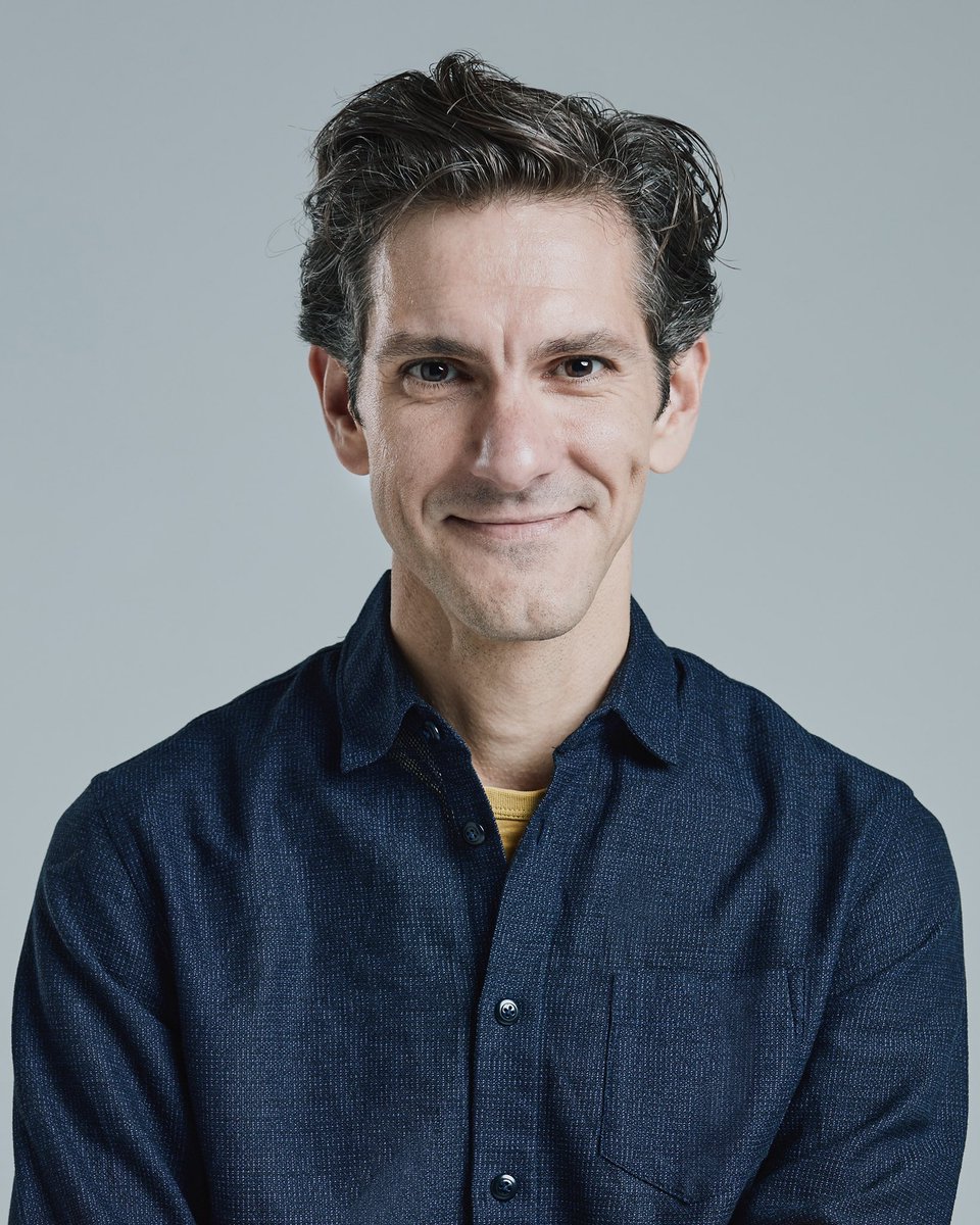 A photo of Mathew Baynton smiling against a grey background.