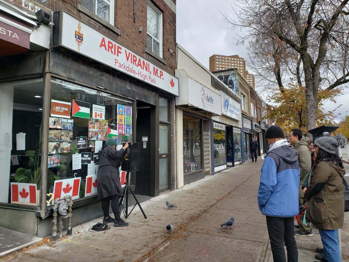 Photo of the outside sidewalk scene. The storefront has a white background and red text saying "Arif Virani, M.P.  Parkdale-High Park." The window has two Palestinian flags newly in the window. A small group gathers on the sidewalk outside.