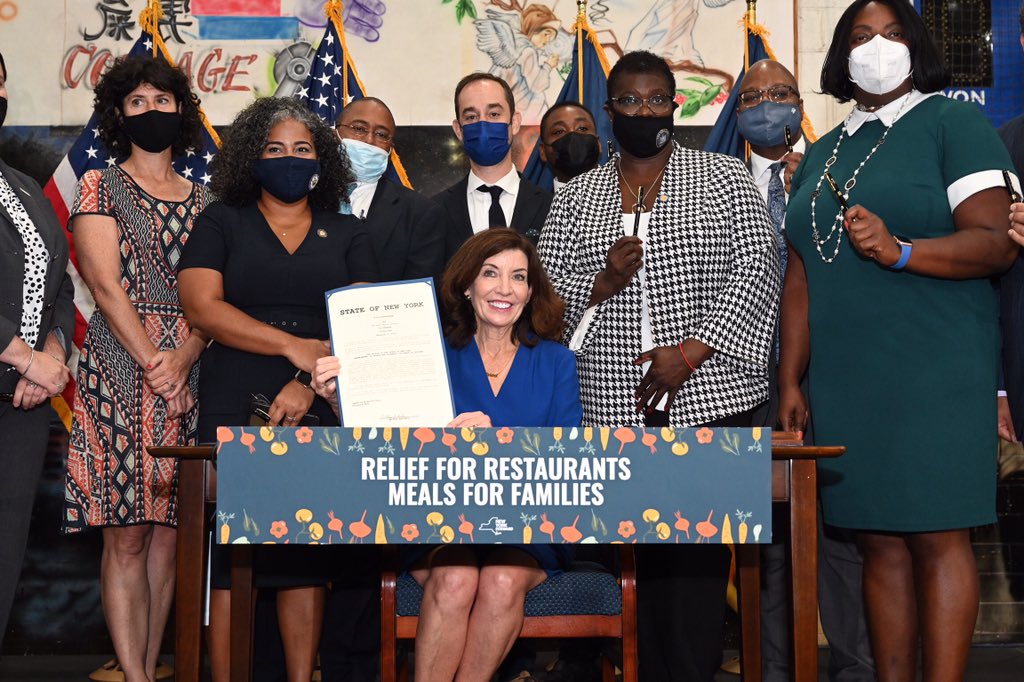 Governor Hochul sits at table holding signed Restaurant Relief Program Act bill surrounded by people