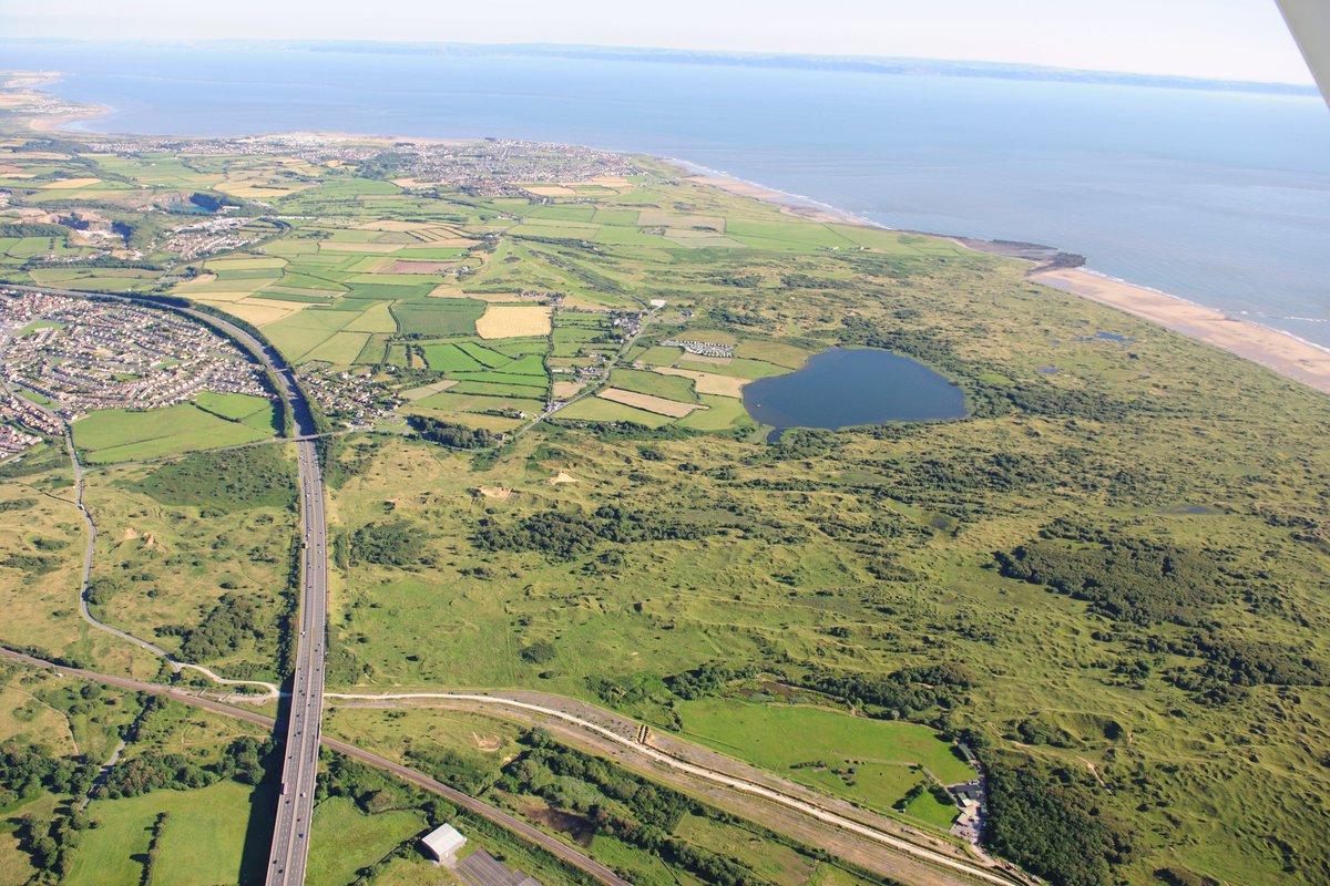 Aerial view showing a fairly flat landscape stretching towards the sea. The boundary between the sea and the land is clearly defined by sand. Near the middle of the photograph is a large lake, whilst on the extreme left is the end of North Cornelly, with the M4 running alongside.