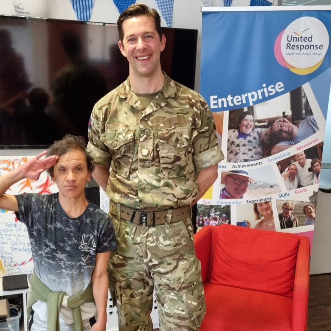 Soldier Tom is stood on the right wearing camouflage. Kayleigh is stood to his left doing a salute. She has short brown curly hair and she is wearing a grey tshirt and a green jumper tied around her waist.