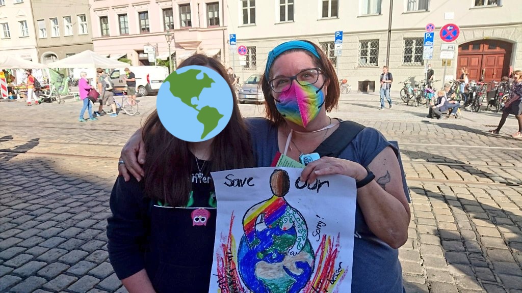 Zwei Frauen Arm in Arm mit Regenbogen Masken und Demoschild auf einen großen Platz der noch recht leer ist.