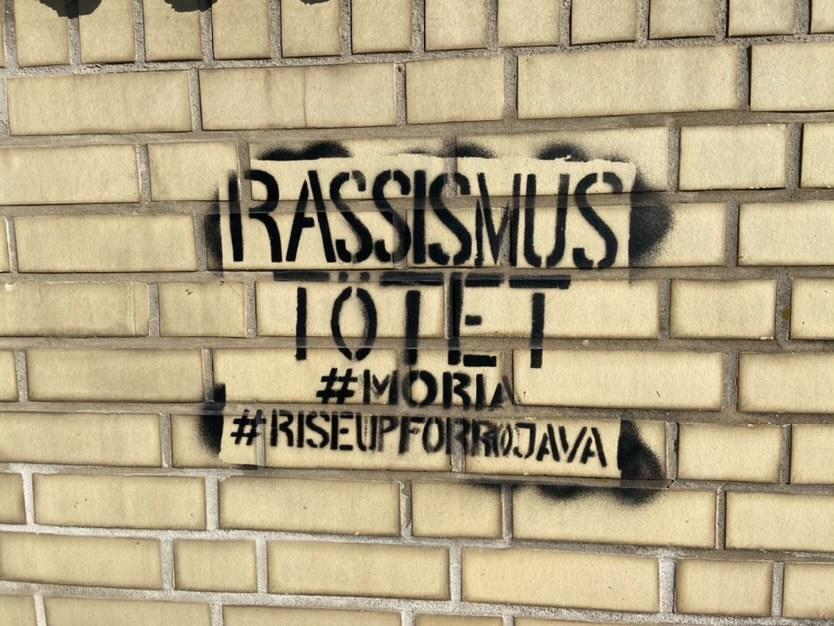 Black stencil on a brick wall that says:
"Racism Kills
#Moria #RiseUpForRojava"