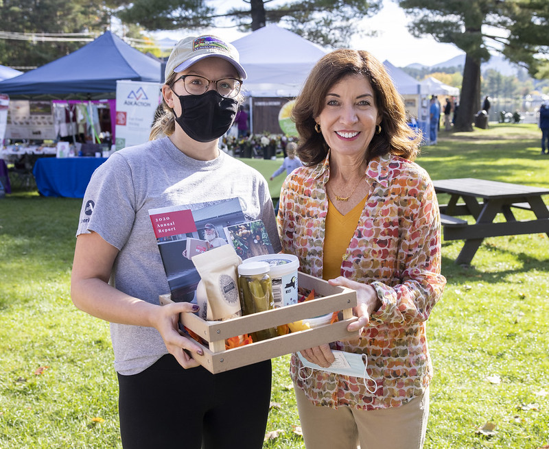 Governor Hochul stand with person holding a small wooden crate with farmers market products at the Saranac Lake Farmers Market
