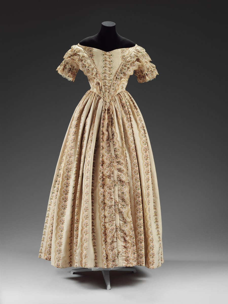 (c)Victoria and Albert Museum, London - A vertical floral printed dress, with the V shaped construction, cap sleeves, and flouce skirt. The color is creme, the print is brown. 