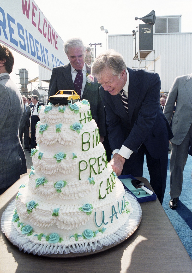 Five layer cake covered in white frosting with white frosting ruffles accented with blue frosting flowers.  There is a yellow toy car on top and it says, on word per layer, top to bottom: Happy Birthday President Carter UAW